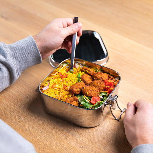 RVS lekvrij lunchbox gevuld met een maaltijd saladen. Een hand zet een vork in de lunchbox